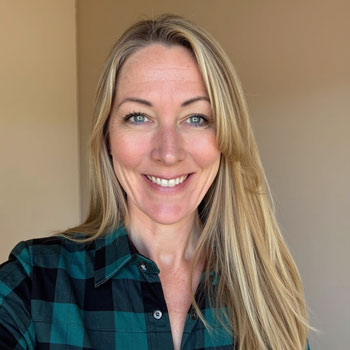 Adult woman with long blond hair standing in front of a tan wall. She's wearing a black and teal plaid shirt.